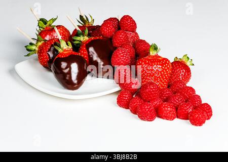 Reichhaltige Makroaufnahme aus in Schokolade getauchten Erdbeeren gepaart mit lebendigen Himbeeren auf weißem Teller; appetitliches Gourmet-Dessert ideal für V Stockfoto