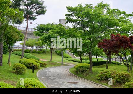 Incheon, Südkorea - 19. Mai 2025: Ein gewundener, gepflasterter Weg führt durch üppige grüne Landschaftsgärten und farbenfrohe Frühlingsbäume in Sicheon Rastplatz, Stockfoto