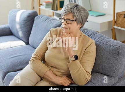 Eine ältere, nachdenkliche Frau mit Brille und bequemer Kleidung, die auf dem Sofa sitzt Stockfoto