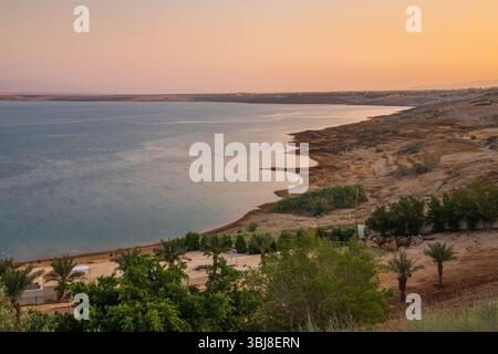 Blick auf den Sonnenuntergang über die Küste des Toten Meeres in Jordanien, mit Palmen, ruhigem Wasser und einer ruhigen Wüstenlandschaft unter goldenem Licht. Stockfoto