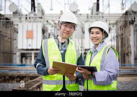 Zwei Ingenieure, die in Hochspannungs-Umspannwerken arbeiten, tragen einen Sicherheitshelm und eine reflektierende Weste und halten das Clipboard und das Tablet mit einem selbstbewussten Lächeln fest Stockfoto