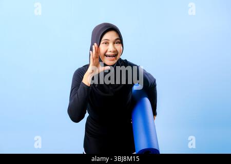 Eine Frau mit schwarzem Kopftuch und blauer Hose beugt sich. Sie macht eine Geste, zu schreien oder etwas anzukündigen. Sie hält ein blaues Yoga Stockfoto