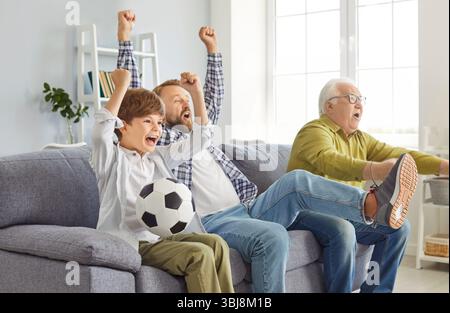 Eine aufregende Familie von Männern aus mehreren Generationen, die auf dem Sofa sitzen und die Lieblingsmannschaft zu Hause anfeuern. Stockfoto