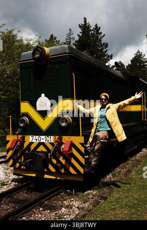 Fröhliche Frau in gelbem Regenmantel mit ausgebreiteten Armen, auf der Vorderseite eines historischen grünen Zuges. Reisekonzept. Mokra gora, Serbien - 17. Mai 2025 Stockfoto