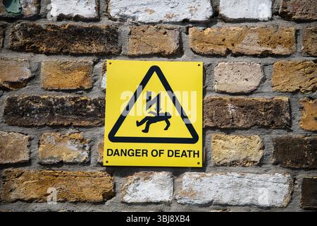 Gelbes Warnschild „Todesgefahr“ an einer Wand mit Blitzschlag und herabfallender Figur, typisch für Sicherheitsschilder in Großbritannien. Stockfoto