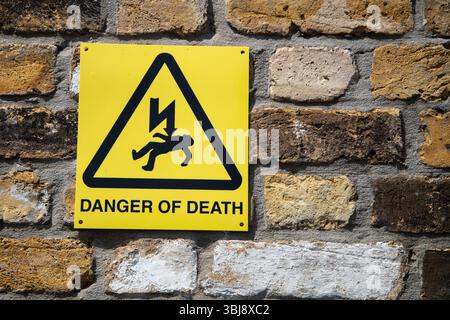Gelbes Warnschild „Todesgefahr“ an einer Wand mit Blitzschlag und herabfallender Figur, typisch für Sicherheitsschilder in Großbritannien. Stockfoto