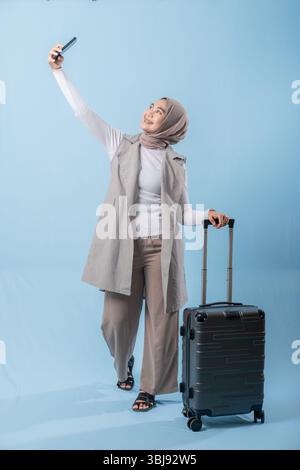 Eine Frau, die einen Schal trägt und ein Handy hält, steht neben einem Koffer. Sie fotografiert sich mit dem Koffer Stockfoto