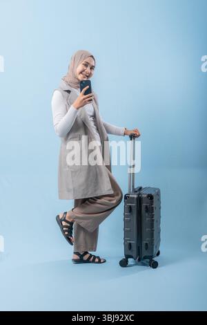 Eine Frau, die einen Schal trägt und ein Handy hält, steht neben einem Koffer. Sie fotografiert sich mit dem Koffer Stockfoto