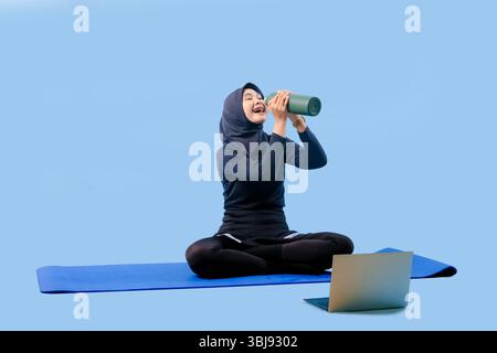 Eine Frau sitzt auf einer Yogamatte mit Laptop und Wasserflasche. Sie lächelt und genießt ihre Zeit Stockfoto
