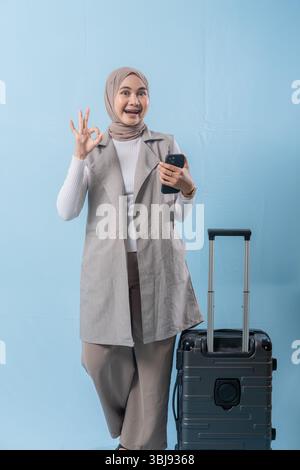 Eine Frau mit Kopftuch hält ein Handy und lächelt. Sie steht neben einem Koffer Stockfoto
