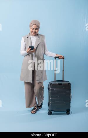 Eine Frau mit Kopftuch hält ein Handy und lächelt. Sie steht neben einem Koffer Stockfoto