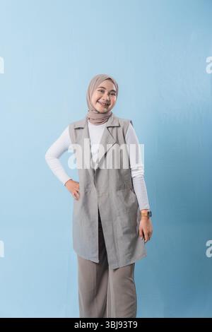 Eine Frau, die eine graue Weste und ein weißes Hemd trägt, lächelt in die Kamera. Sie trägt ein Kopftuch und hat die Arme gekreuzt Stockfoto