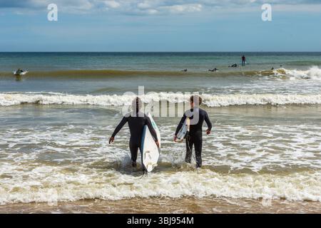 East Cliff Beach, Bournemouth, Großbritannien - 28. Mai 2025: Zwei Surfer gehen mit Surfbrettern ins Meer. Stockfoto