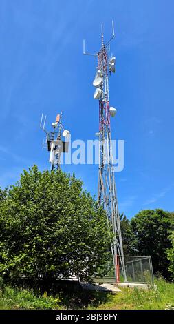 Zwei hohe Metallkommunikationstürme mit mehreren Antennen und Satellitenschüsseln erheben sich über grünen Bäumen und einem umzäunten Bereich, der sich gegen einen klaren Raum befindet Stockfoto
