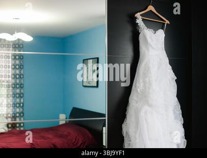 Ein wunderschönes Hochzeitskleid hängt an einem hölzernen Kleiderbügel in einem hell beleuchteten Raum mit blauen Wänden und einem gemütlichen Bett. Stockfoto