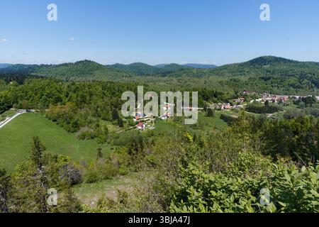 Ein malerisches Dorf eingebettet zwischen sanften, bewaldeten Hügeln unter einem klaren blauen Himmel, mit lebhaften grünen Wiesen im Vordergrund. Stockfoto