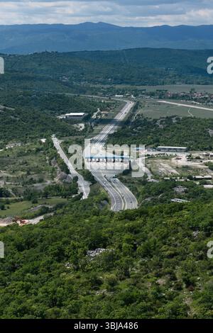 Ein erhöhter Blick fängt eine Autobahn ein, die sich durch ein üppiges, grünes Tal schlängelt, mit einer Mautstelle, und entfernten Bergen unter einem bewölkten Himmel Stockfoto