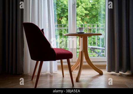 Eine gemütliche und ruhige Szene mit einem roten Stuhl und einem runden Holztisch mit einer Tasse Kaffee, neben einem großen Fenster mit transparenten Vorhängen. Stockfoto