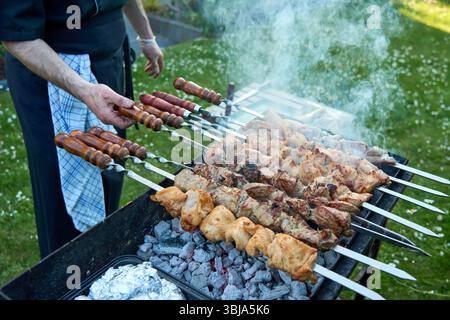 Mann auf Schürze bereitet verschiedene Fleischspieße auf einem rauchenden Holzkohlegrill in einer Gartenanlage zu. Stockfoto