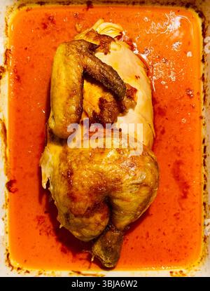 Gebratenes Hähnchen in scharfer Roter Sauce. Blick von oben auf ein ganzes geröstetes Huhn, serviert in einer tiefen Auflaufform, gefüllt mit lebendiger roter Sauce. Stockfoto