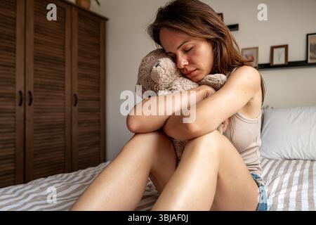 Traurige junge Frau, die im Zimmer auf dem Bett sitzt und Teddybären umarmt Stockfoto