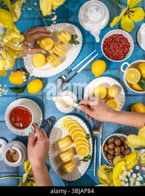 Zwei Frauen teilen sich eine herrliche Teezeit mit Zitronenmakronen, Tee, geschnittenen Zitronen und verschiedenen Leckereien auf einem blau gekachelten Tisch mit gelben Tulpen Stockfoto