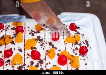 Cremiger, geschichteter Kuchen mit Stücken Schokolade, Kirsch und Pfirsich, geschnitten mit einem großen Küchenmesser, Nahaufnahme. Stockfoto