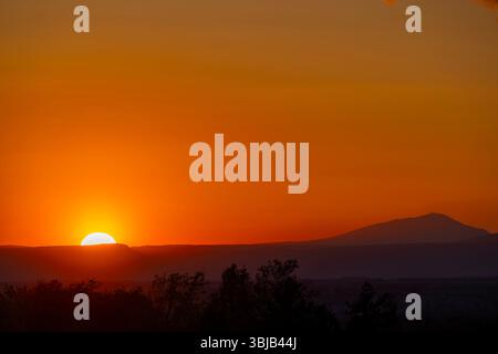Die Sonne untergeht hinter den Bergen und schafft Silhouetten von Bäumen mit orangefarbenem Himmel Stockfoto