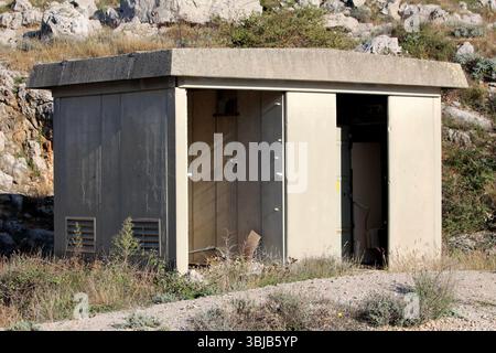 Kleiner Betonschuppen mit flachem Dach und offener Metalltür steht auf felsigem Boden mit trockenem Gras und Sträuchern, die von Kalksteinfelsen unterlegt sind Stockfoto