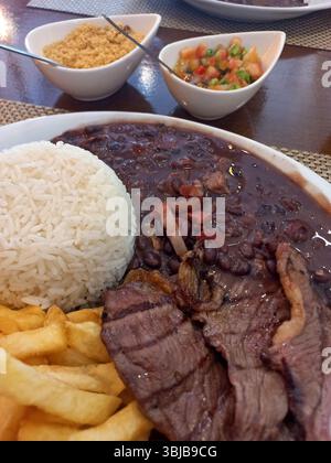 Typisch brasilianische Mahlzeit mit weißem Reis, schwarzen Bohnen, gegrilltem Rindfleisch picanha, Pommes, Vinaigrette, Salsa und Farofa, serviert auf einem Restauranttteller Stockfoto