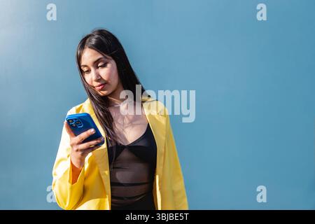 Selbstbewusste junge Frau in einem stilvollen gelben Blazer und einem transparenten schwarzen Oberteil schaut auf ihr Handy, während sie bei hellem Tageslicht vor einer glatten blauen Wand steht. Stockfoto