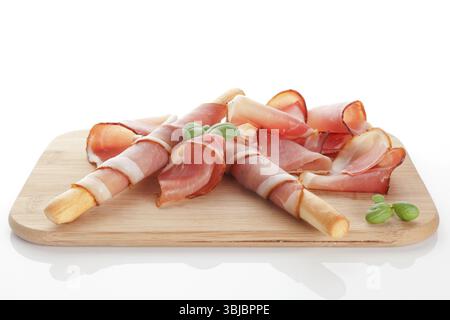 Prosciutto-Scheiben mit Brotstab und frischem Basilikum auf hölzernem Schneidebrett isoliert auf weißem Hintergrund. Kulinarischer italienischer Schinken Stockfoto