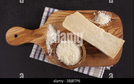 Geriebenes und ganzes Stück Parmesankäse auf Holzbrett auf schwarzer Oberfläche von oben Stockfoto