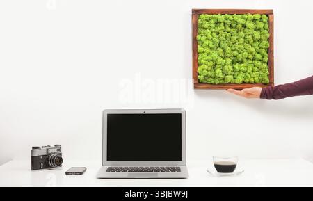 Minimalistischer geschäftlicher Arbeitsplatz mit Laptop, Mobiltelefon, Kaffee isoliert auf weißem Hintergrund. Weibliche Hand, die Moosmalerei hält. Modern zeitgenössisch Stockfoto