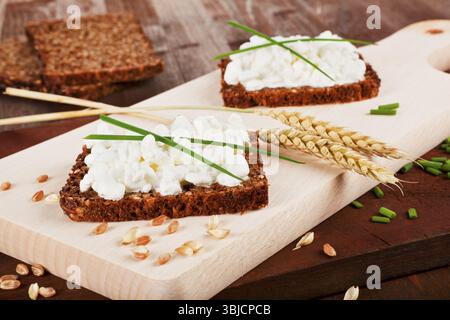 Schwarze Vollkornbrotscheiben mit Hüttenkäse, frischen Kräutern und Weizen auf Holzschneidebrett. Gesunde rustikale Ernährung Hintergrund Stockfoto