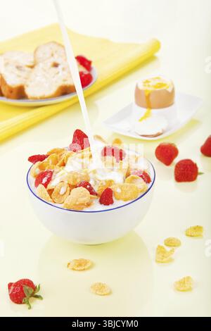 Gießen Sie Milch in eine Schüssel mit Cornflakes und Erdbeeren. Leckeres Frühstück. Cornflakes mit Erdbeeren in der Schüssel, Brot und Ei im Hintergrund Stockfoto