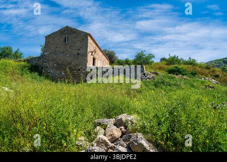 Kleines Steingebäude auf einem grasbewachsenen Hügel bei Akrokorinthos, umgeben von Ruinen und wildem Grün unter einem hellen, ruhigen Himmel. Stockfoto
