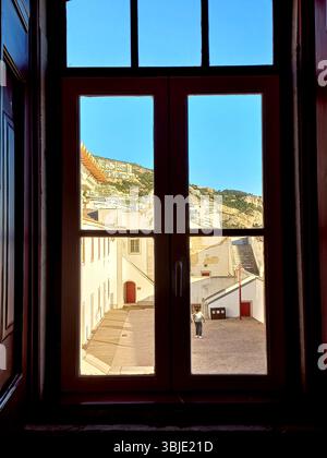 Sesimbra, Portugal. 20. Dezember 2024 Ein dunkel gerahmtes Fenster bietet einen gerahmten Blick auf den weiß getünchten Innenhof von Sesimbra, die Gebäude an den Küsten und Go Stockfoto