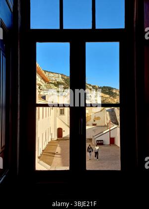 Sesimbra, Portugal. 20. Dezember 2024 Ein dunkel gerahmtes Fenster bietet einen gerahmten Blick auf den weiß getünchten Innenhof von Sesimbra, die Gebäude an den Küsten und Go Stockfoto