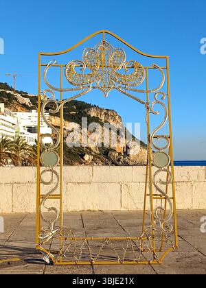 Sesimbra, Portugal. 20. Dezember 2024 Ein goldener Metallrahmen steht leer vor Küstenklippen und azurblauer Meereskulisse, flankiert von Palmen Stockfoto