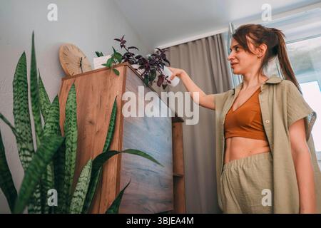 Frau mit gebundenen braunen Haarspray Zimmerpflanzen von hinten in weich beleuchtetem Raum mit Holzschrank, Topfblumen, Home Wellness, Indoor Gartenarbeit Stockfoto