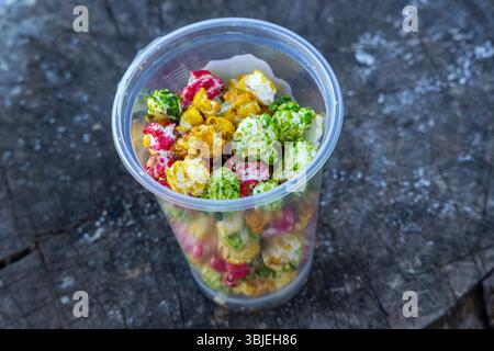 Köstliches buntes süßes Popcorn in einem klaren Plastikbecher. Strukturierter Holzhintergrund. Kinosnack, Süßigkeiten, Leckereien. Stockfoto