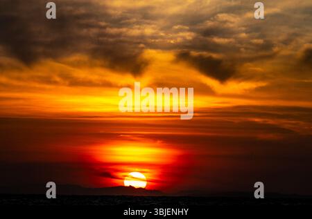 Ein atemberaubender Sonnenuntergang über dem Meer mit leuchtenden Orange-, Gelb- und Rottönen, die sich in einem dramatischen Himmel voller Wolken verschmelzen. Stockfoto