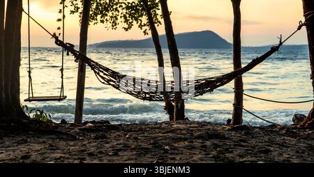 Eine ruhige Strandszene mit einer Hängematte und einer Schaukel zwischen Bäumen, mit sanften Wellen am Ufer und einer fernen Insel bei Sonnenuntergang Stockfoto