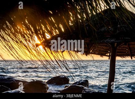 Eine ruhige Strandszene bei Sonnenuntergang mit einem strohgedeckten Schirm vor dem leuchtenden orange-gelben Himmel. Wellen krachen sanft auf die Felsen Stockfoto