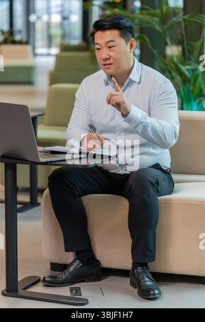 Junger chinesischer Geschäftsmann, der sich in der Hotellobby auf einem Laptop aufschreibt oder Notizen macht, während er an einem Online-Videokurs oder einem Büromeeting teilnimmt. Selbstbewusster Manager im Bereich T-Shirt-Support, der Webcam ansieht Stockfoto