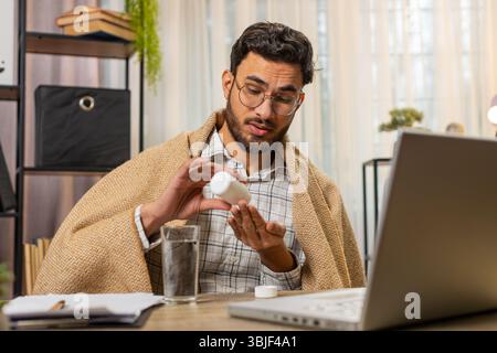 Gesundheit, Kälte und Menschen Konzepte. Der kranke junge arabische Geschäftsmann, der in eine Decke gehüllt ist, arbeitet am Laptop am Schreibtisch im Heimbüro. Ungesunder Hispanic Freelancer nimmt Medikamente und zittert vor der Kälte zu Hause Stockfoto