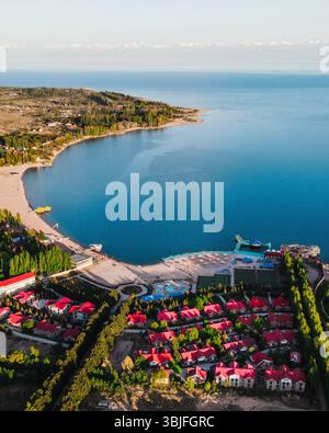 Weite Luftaufnahme des Issyk Kul Lake, der sein riesiges blaues Wasser, eine gekrümmte Küste, ein Resort mit roten Häusern und eine üppig grüne Halbinsel enthüllt Stockfoto