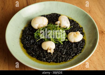 Ein Gourmet-Teller mit Risotto aus schwarzer Tintenfisch-Tinte, garniert mit gebratenem Tintenfisch und einer frischen Rucola-Garnitur. Der leuchtende Grün- und Tiefschwarz-Kontrast b Stockfoto