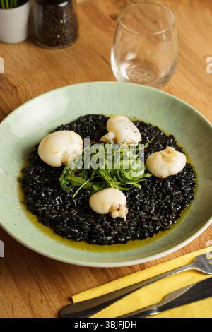 Ein Gourmet-Teller mit Risotto aus schwarzer Tintenfisch-Tinte, garniert mit gebratenem Tintenfisch und einer frischen Rucola-Garnitur. Der leuchtende Grün- und Tiefschwarz-Kontrast b Stockfoto